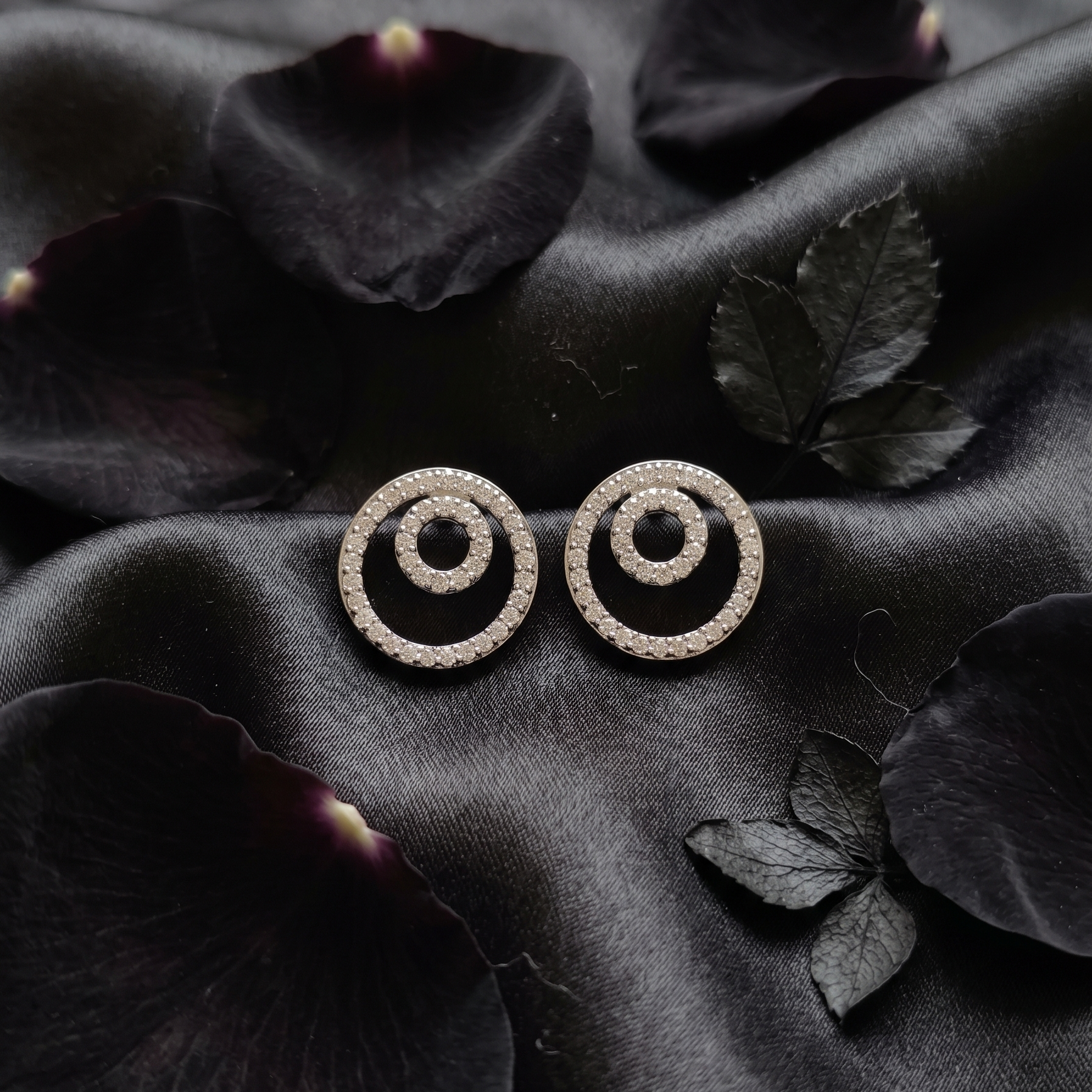 Pair of circular earrings on a dark fabric background with leaves.