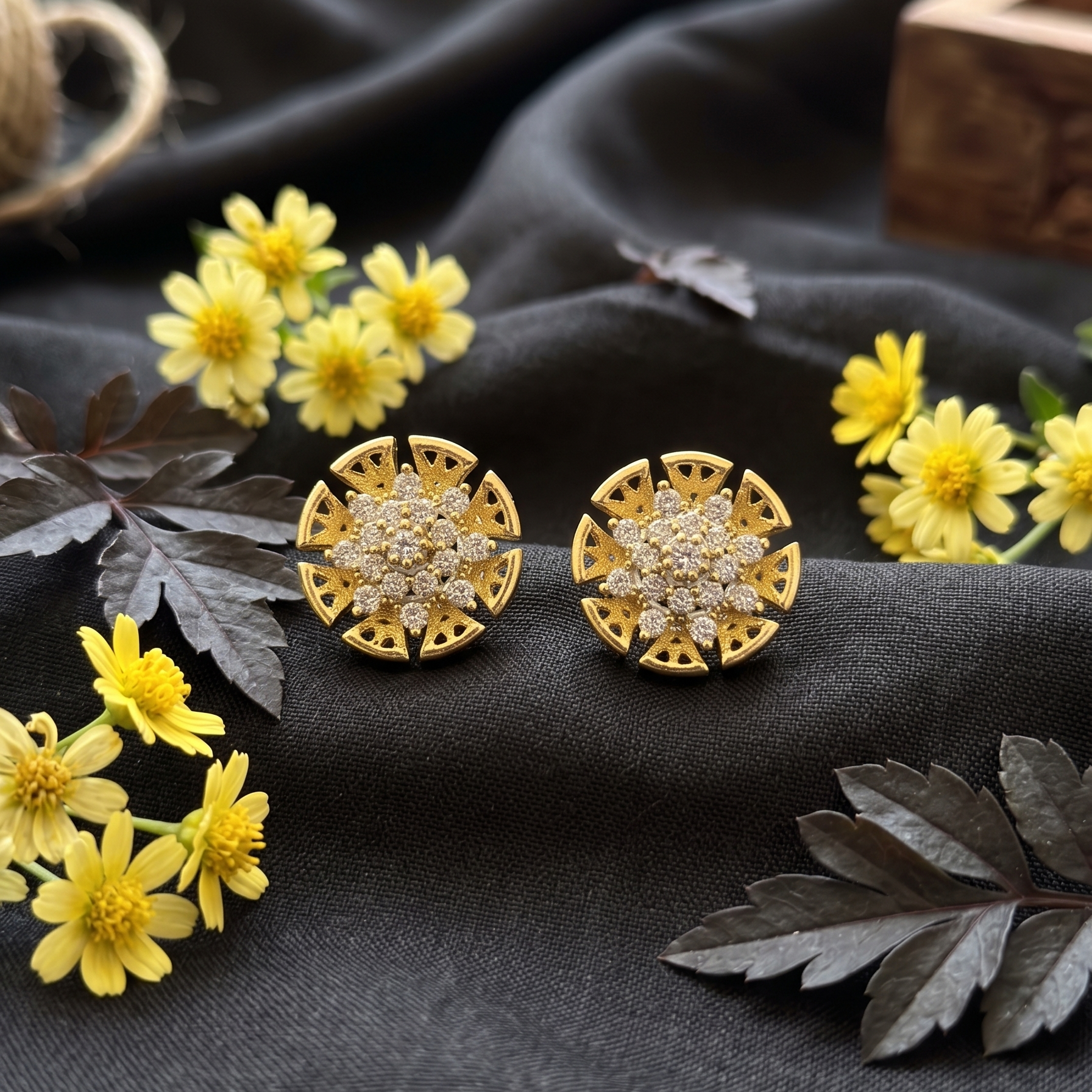 Gold earrings with diamond accents on a dark fabric background with yellow flowers and leaves.