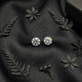 Pair of silver stud earrings on black fabric with decorative leaves.