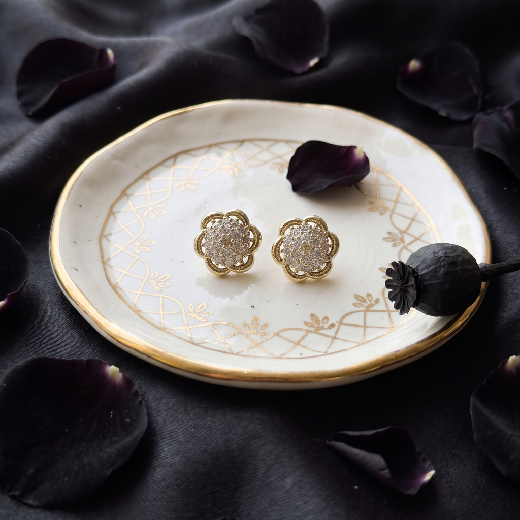 Gold earrings on a decorative plate with black fabric and purple flowers in the background
